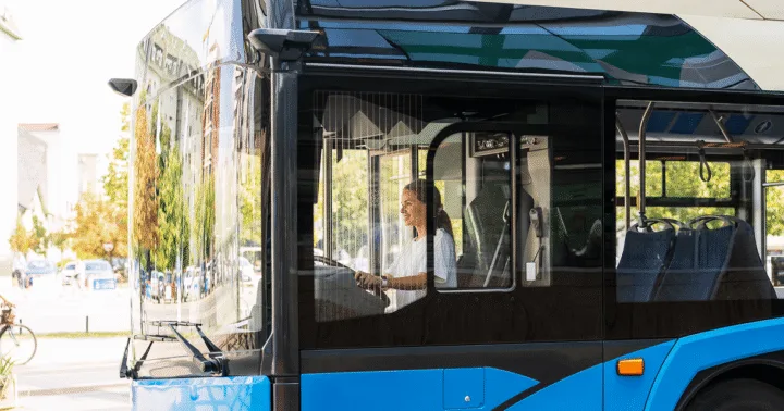 Motorista ao volante de ônibus urbano azul parado em rua arborizada, vista lateral da cabine