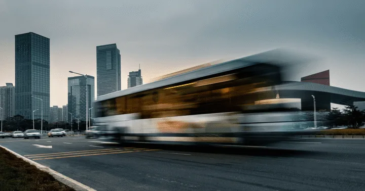 Ônibus urbano em movimento numa avenida ao anoitecer, com arranha-céus da cidade ao fundo