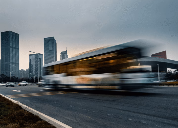 Ônibus urbano em movimento numa avenida ao anoitecer, com arranha-céus da cidade ao fundo