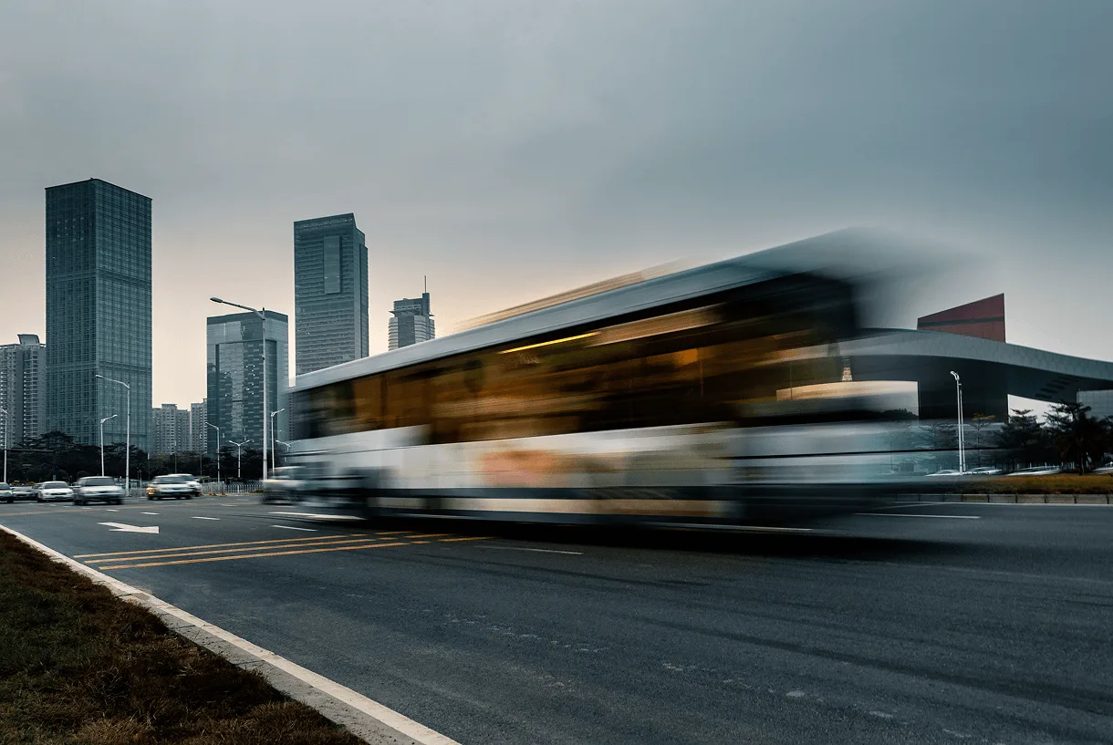 Ônibus urbano em movimento numa avenida ao anoitecer, com arranha-céus da cidade ao fundo