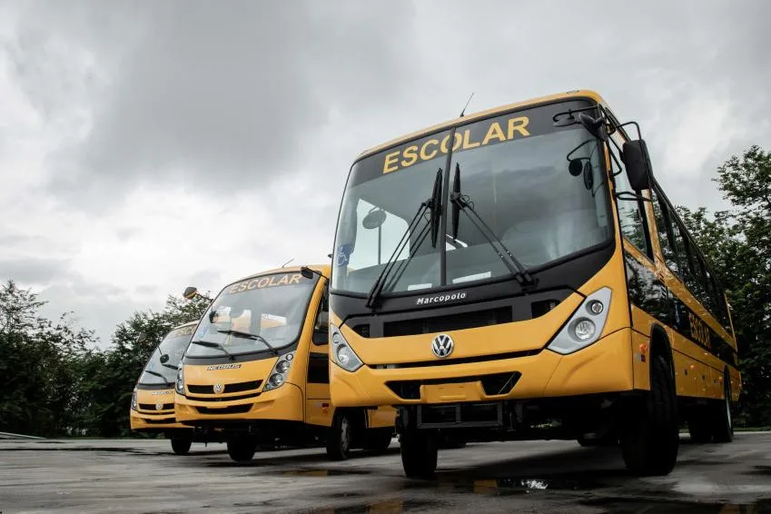 Três ônibus escolares amarelos com emblema Volkswagen, em fila e vistos de frente, em pátio sob céu nublado