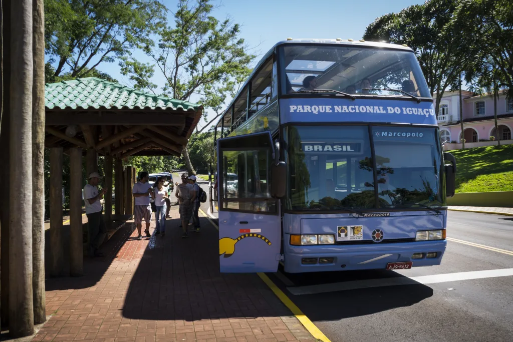Ônibus de turismo Marcopolo de dois andares, azul-claro, parado no Parque Nacional do Iguaçu com turistas ao fundo