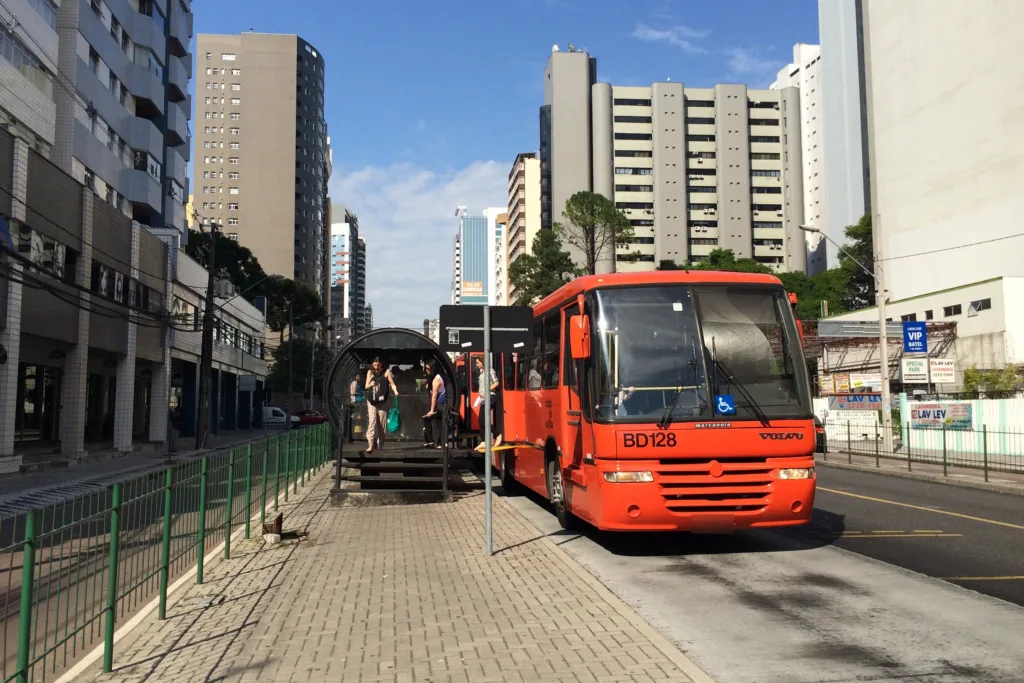 Ônibus laranja da frota de Curitiba chegando a uma estação-tubo, com prédios ao fundo