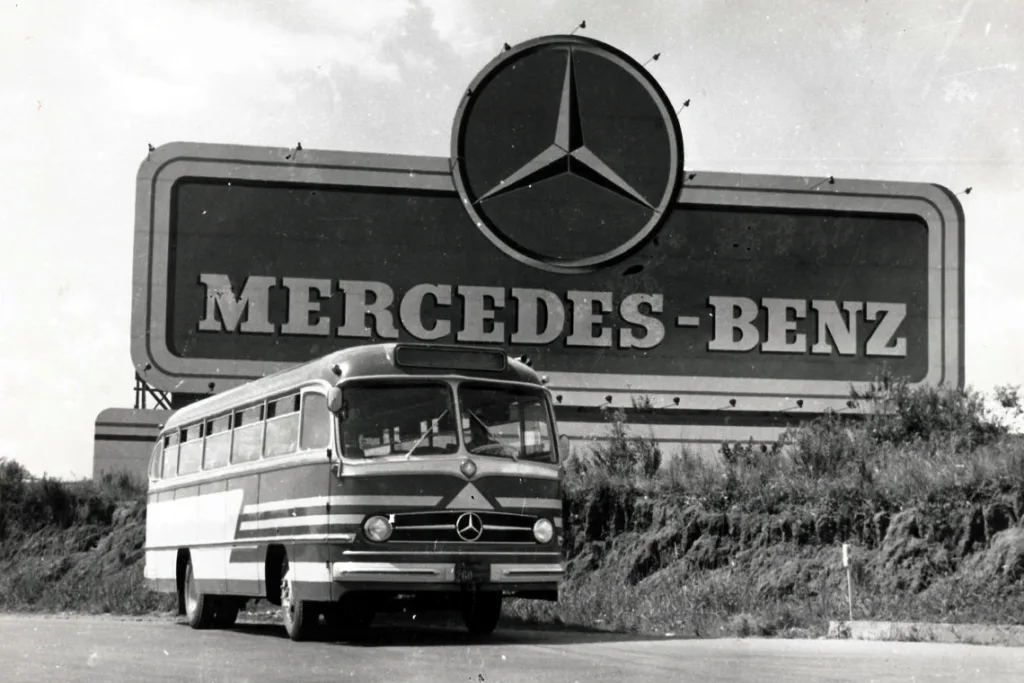 Fotografia histórica em preto e branco de um ônibus Mercedes-Benz dos anos 1950 diante de um grande letreiro da marca com a estrela