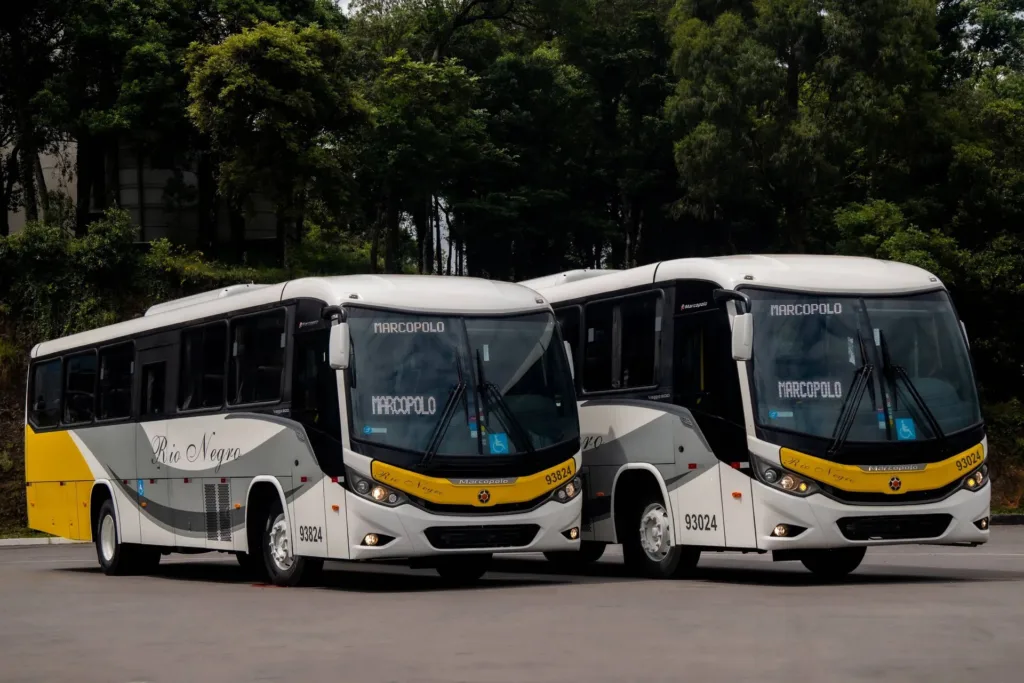 Dois ônibus Marcopolo da Rio Negro Transportes, brancos com detalhes em amarelo e cinza, estacionados lado a lado em pátio cercado de árvores