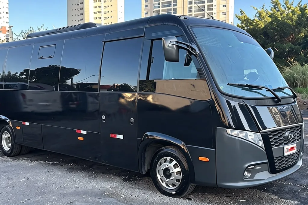 Micro-ônibus Marcopolo Volare Fly 9 2024 preto, visto em três quartos dianteiro, num pátio de cascalho com prédios ao fundo