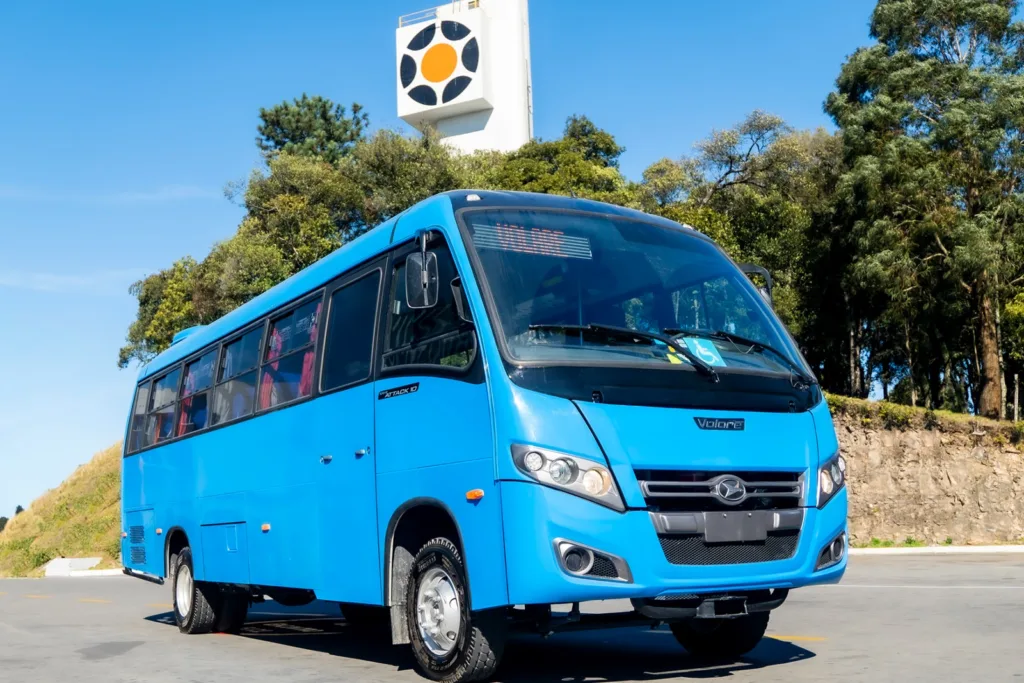 Micro-ônibus Volare Attack 10 Híbrido azul-claro, visto de frente em três quartos, em pátio ensolarado