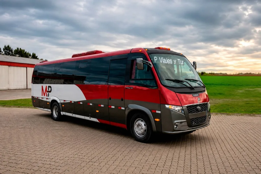 Micro-ônibus Volare Fly 10 Executivo vermelho e cinza da MyP Viajes y Turismo, visto de frente em três quartos, em pátio de pedra com campo ao fundo